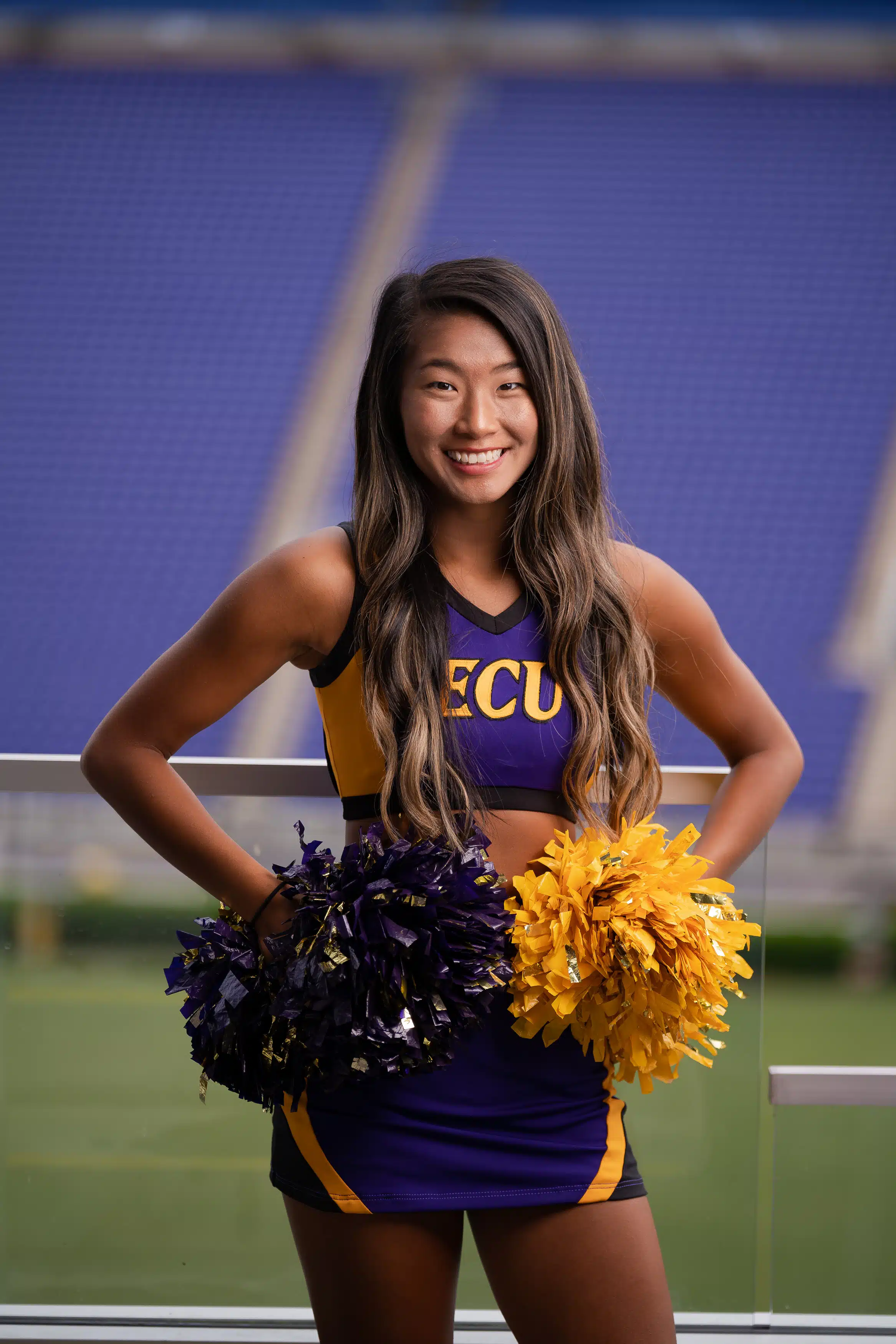 ECU cheerleader posing at dowdy-ficklen stadium for a headshot while wearing a purple and gold cheerleading outfit