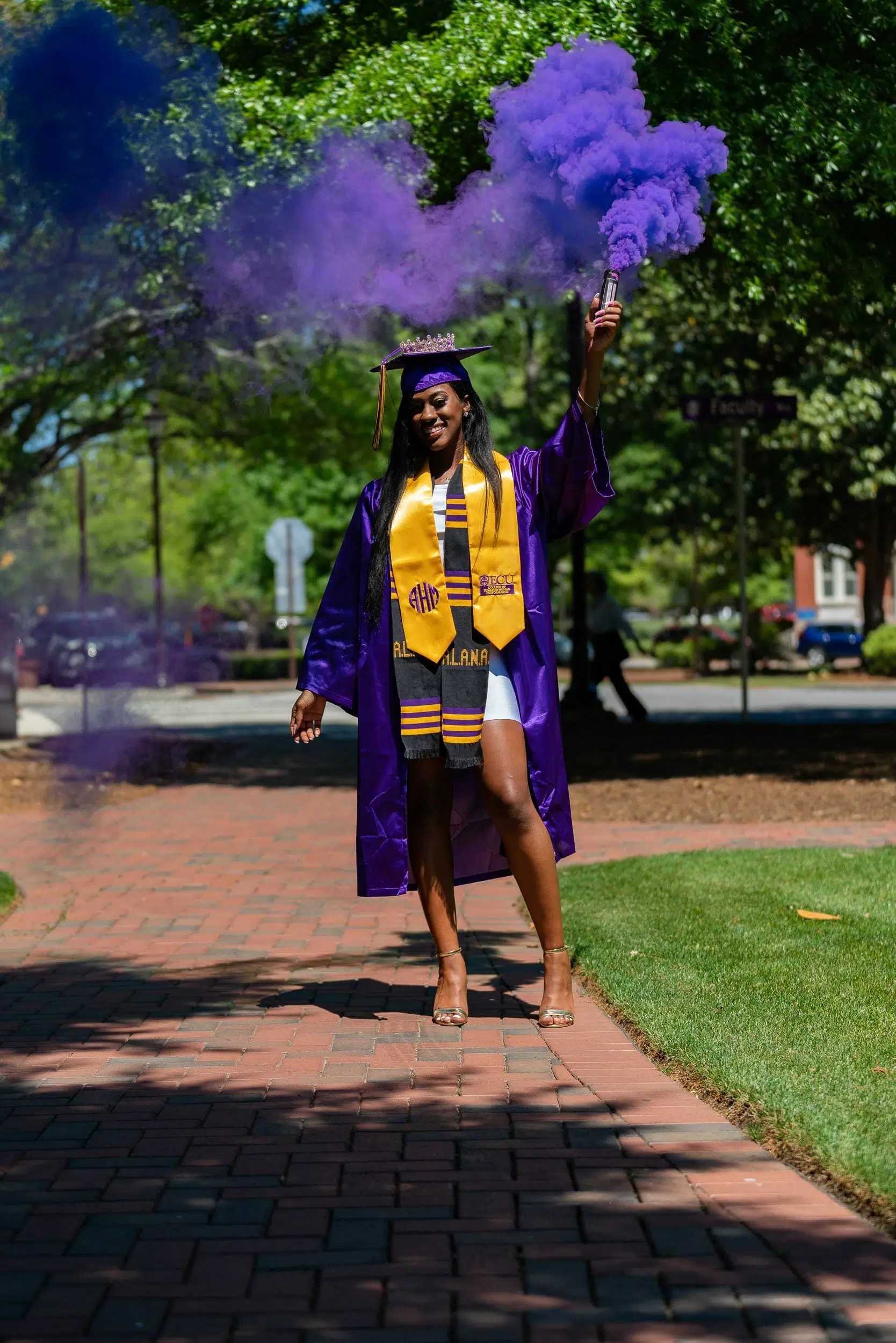 Woman wearing her cap and gown for East Carolina University (ECU) and posing for her graduation photos, while also using a purple smoke bomb.
